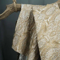 The Luxury Embossed Brocade Fabric by Longan Craft, a premium metallic jacquard with intricate paisley and floral designs in gold and silver, is draped elegantly over a rustic wooden branch tied with rope.