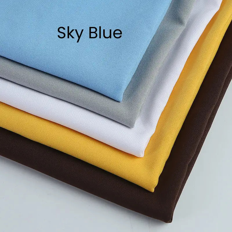 Five neatly folded Longan Craft Matte Finish Gabardine Fabric pieces (sky blue labeled, gray, white, yellow, dark brown) with twill weave are stacked diagonally on a plain surface. Perfect for jackets and wrinkle resistant.