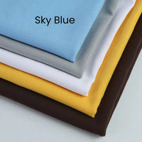 Five neatly folded Longan Craft Matte Finish Gabardine Fabric pieces (sky blue labeled, gray, white, yellow, dark brown) with twill weave are stacked diagonally on a plain surface. Perfect for jackets and wrinkle resistant.