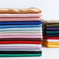 A neatly stacked pile of Longan Craft Matte Satin Fabric By The Yard in various solid colors—perfect for bridesmaid and wedding dresses—with a smaller stack of multicolored satin textiles in the background.