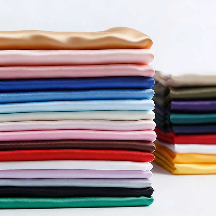 A neatly stacked pile of Longan Craft Matte Satin Fabric By The Yard in various solid colors—perfect for bridesmaid and wedding dresses—with a smaller stack of multicolored satin textiles in the background.