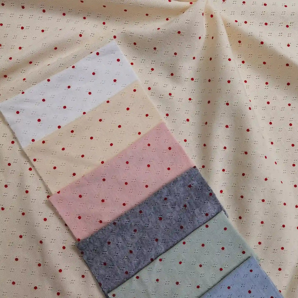 A stack of Mini Red Dot Printed Pointelle Fabric By The Yard swatches in white, cream, pink, grey, green, and blue, all with a small red dot print, layered over a larger piece of the same cream cotton jersey fabric.