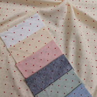 A stack of Mini Red Dot Printed Pointelle Fabric By The Yard swatches in white, cream, pink, grey, green, and blue, all with a small red dot print, layered over a larger piece of the same cream cotton jersey fabric.