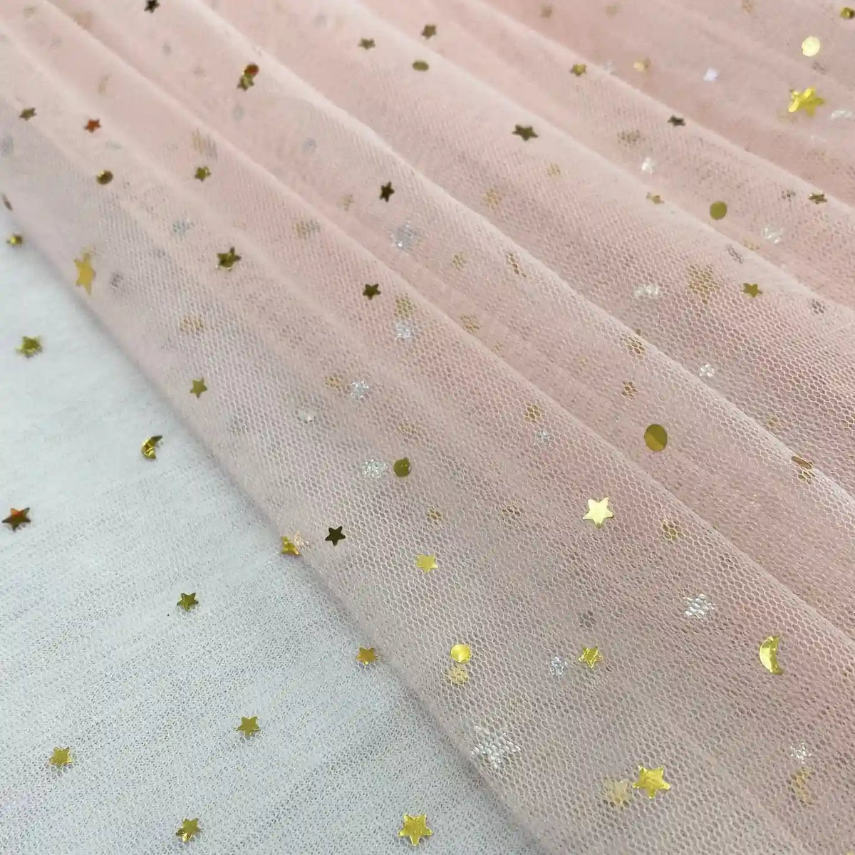 A close-up of Longan Craft’s Moon Star Sequins Tulle Fabric by the yard—multicolor glitter tulle, ideal for stage costumes—shows shiny gold star and dot sequins on pale pink tulle, laid over a white surface with gold stars.