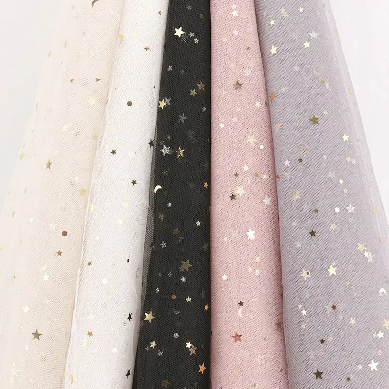 Five Moon Star Sequins Tulle Fabrics by Longan Craft in cream, white, black, pink, and lavender hang side by side. Each multicolor glitter tulle is ideal for stage costumes or elegant attire.