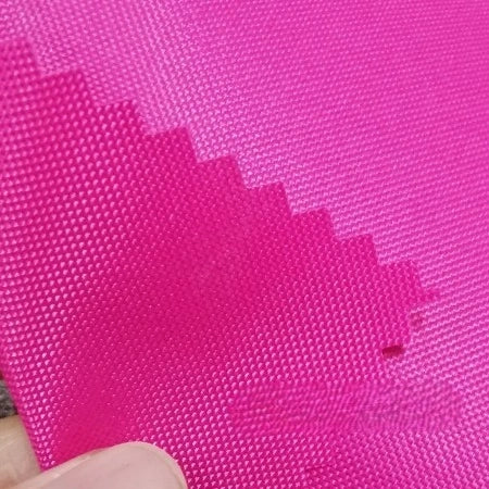 A close-up of two bright pink pieces of Longan Craft Multicolor 300D Oxford Fabric with zigzag edges; a person’s fingers hold the water-repellent, PU-coated polyester layers made for bags and backpacks.