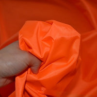 A hand grips a piece of bright orange Longan Craft Multicolor 300D Oxford Fabric by the yard, PU coated polyester ideal for bags and backpacks, creating folds in the durable, water repellent material with the same fabric in the background.