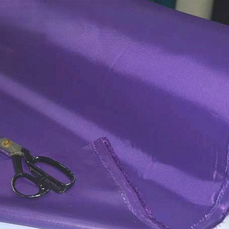 A pair of black-handled scissors rests on a piece of shiny purple Multicolor 300D Oxford Fabric by the yard from Longan Craft, a durable PU coated polyester material perfect for bags and backpacks, with a partially cut edge.
