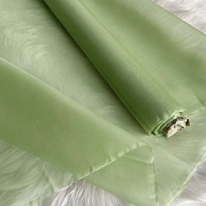 A roll of light green, sheer organza lies on a soft white surface. This smooth, slightly translucent Longan Craft Multicolor 8 Momme Organza Silk Fabric is perfect for evening gowns or pairing with luxury pure mulberry silk material.