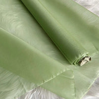 A roll of light green, sheer organza lies on a soft white surface. This smooth, slightly translucent Longan Craft Multicolor 8 Momme Organza Silk Fabric is perfect for evening gowns or pairing with luxury pure mulberry silk material.