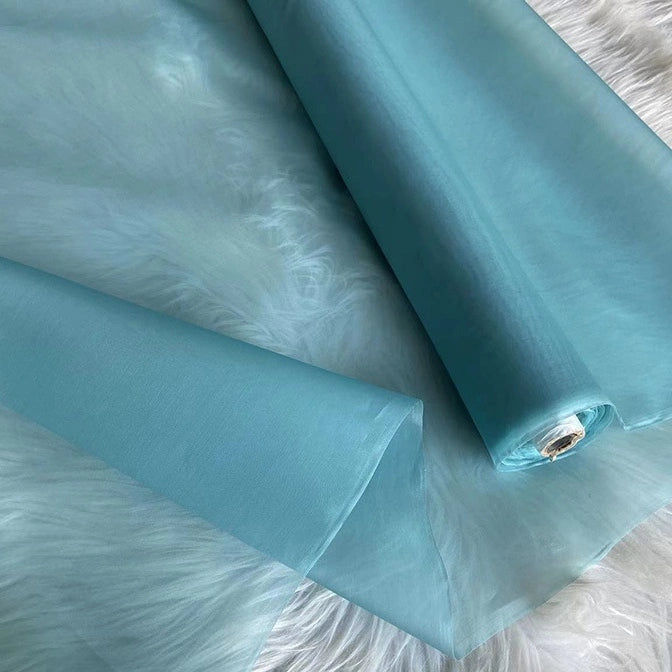A roll of Longan Craft's Multicolor 8 Momme Organza Silk Fabric By The Yard, made from pure mulberry silk, is partly unrolled on a white furry surface. Its delicate, semi-transparent luxury is ideal for elegant dresses and evening gowns.