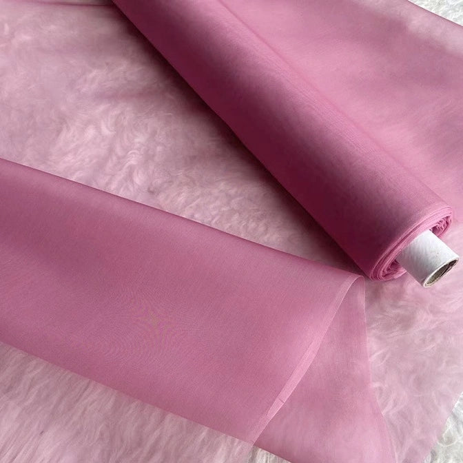 A roll of Longan Craft Multicolor 8 Momme Organza Silk Fabric By The Yard, a pure mulberry silk material for luxury dresses, is partially unrolled on a light pink textured surface, showing its smooth and delicate translucent appearance.