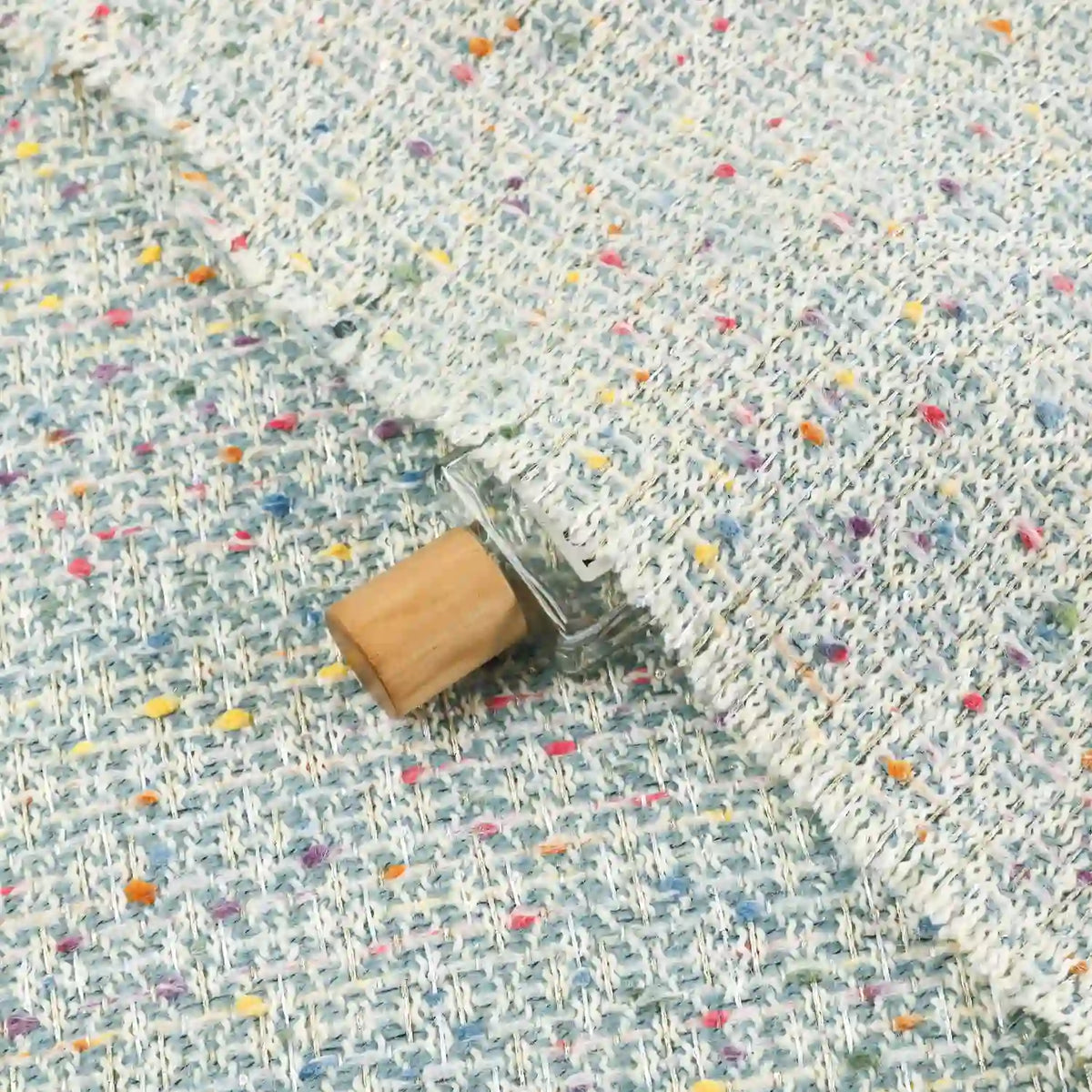 A small glass vial with a wooden cap is partially tucked under Longan Craft’s Multicolor Dot Boucle Tweed Fabric— a heavyweight jacquard wool blend suiting material— its vibrant, multicolored specks adding playful texture to the cozy scene.