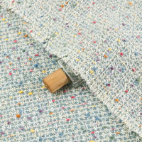 A small glass vial with a wooden cap is partially tucked under Longan Craft’s Multicolor Dot Boucle Tweed Fabric— a heavyweight jacquard wool blend suiting material— its vibrant, multicolored specks adding playful texture to the cozy scene.