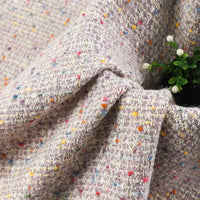A close-up of the Multicolor Dot Boucle Tweed Fabric by Longan Craft showcases its chic heavyweight jacquard wool blend, featuring a light base with colorful specks and elegant soft folds. A small green plant with white flowers peeks out from between the draped suiting fabric.
