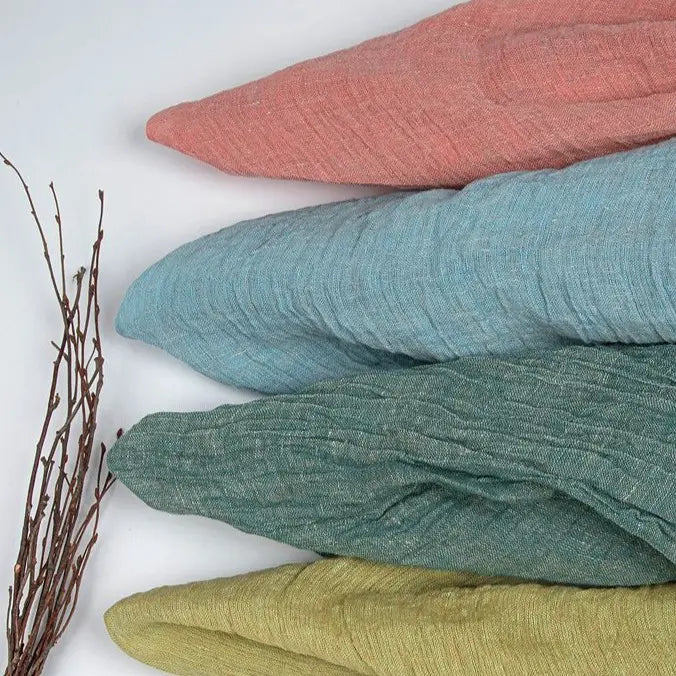 Four pieces of Longan Craft’s Multicolor Heathered Crepe Fabric by the Yard, made from 100% linen sandwashed crepe material in pink, blue, green, and yellow shades, are folded and stacked diagonally. To the left of the stack on a light surface lies a small bundle of thin brown twigs.