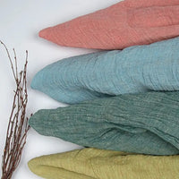 Four pieces of Longan Craft’s Multicolor Heathered Crepe Fabric by the Yard, made from 100% linen sandwashed crepe material in pink, blue, green, and yellow shades, are folded and stacked diagonally. To the left of the stack on a light surface lies a small bundle of thin brown twigs.