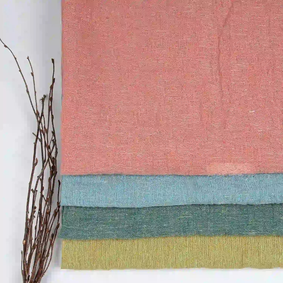 Four folded pieces of Longan Craft’s Multicolor Heathered Crepe Fabric By The Yard—100% linen sandwashed crepe in pink, blue, green, and yellow—are stacked on a white surface. A small bundle of brown twigs rests beside them on the left.