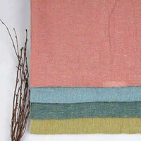 Four folded pieces of Longan Craft’s Multicolor Heathered Crepe Fabric By The Yard—100% linen sandwashed crepe in pink, blue, green, and yellow—are stacked on a white surface. A small bundle of brown twigs rests beside them on the left.