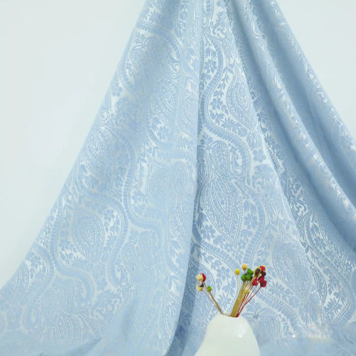 The Multicolor Jacquard Damask Fabric by Longan Craft, featuring a floral cotton damask pattern, is draped against a pale wall while a white vase of vibrant dried flowers sits in the foreground.