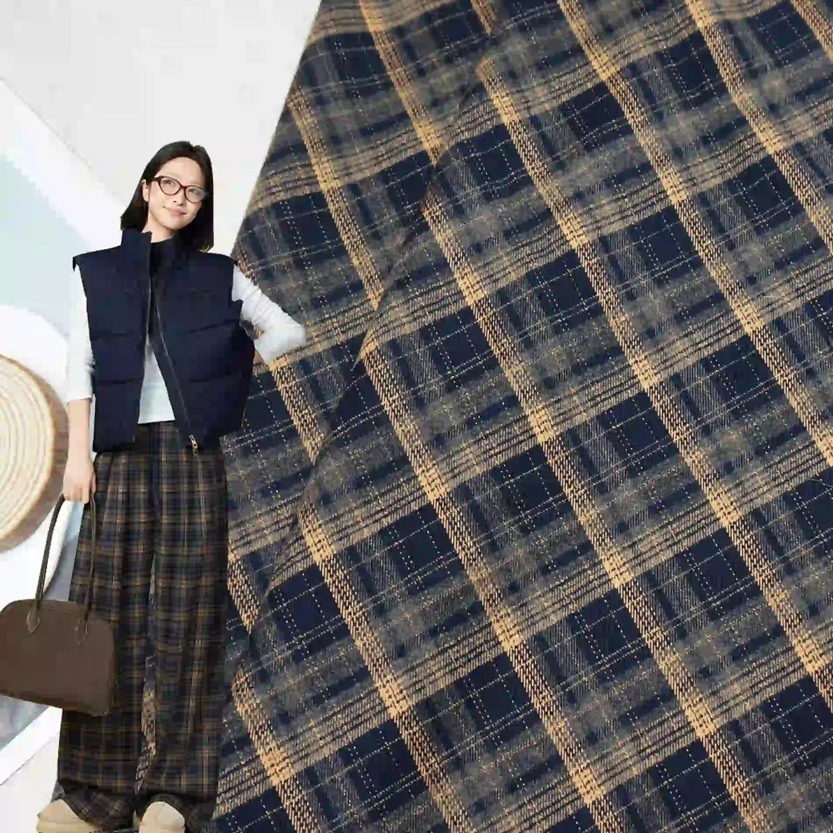 A woman wearing glasses, a navy vest, and wide-leg plaid pants holds a brown bag. The background displays Longan Craft Multicolor Mammoth Plaid Brushed Flannel Fabric, perfect for cozy pants.