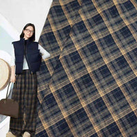 A woman wearing glasses, a navy vest, and wide-leg plaid pants holds a brown bag. The background displays Longan Craft Multicolor Mammoth Plaid Brushed Flannel Fabric, perfect for cozy pants.