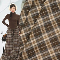 A woman in a brown turtleneck, plaid wide-leg pants, and earmuffs stands before Longan Craft’s Multicolor Mammoth Flannel Fabric By The Yard—plaid brushed flannel textile perfect for cozy pants.