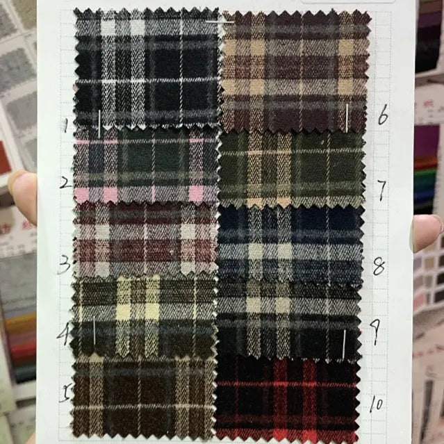 A hand displays a chart with ten numbered plaid swatches on white grid paper, featuring the Multicolor Mammoth Flannel Fabric By The Yard from Longan Craft. Each swatch shows unique checkered patterns and vibrant colors ideal for cozy pants.