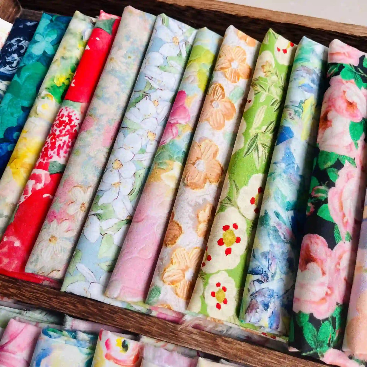 A wooden tray displays neatly rolled Multicolor Printed Lawn Fabric By The Yard from Longan Craft, featuring soft floral cotton lawn fabric in various pastel colors and patterns, arranged side by side.