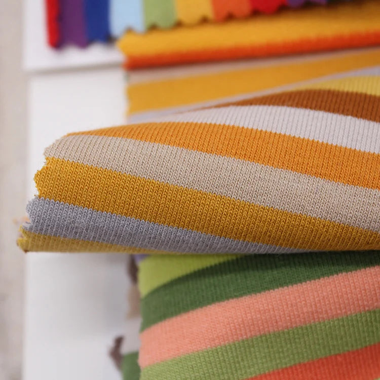 A close-up of Longan Craft's Multicolor Striped Cotton French Terry Fabric by the yard, featuring yellow, white, and gray stripes in front, with other bright striped stretch knit French terry materials for sweatshirts in the background.