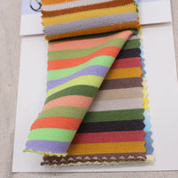 A sample card displays several swatches of Longan Craft’s Multicolor Striped Cotton French Terry Fabric by the yard, featuring horizontal stripes in orange, green, yellow, purple, brown, and blue on a white background.