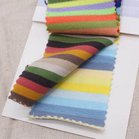 A sample book of Longan Craft Multicolor Striped Cotton French Terry Fabric swatches, featuring bright and pastel horizontal stripes, is displayed on a beige surface. Stretch knit by the yard for warm sweatshirts.