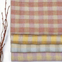 Four folded pieces of Longan Craft’s Multicolor Washed Plaid Linen Fabric By The Yard, featuring vintage checkered pastel patterns in pink, yellow, and blue tones, are stacked beside a twig with thin branches on a white background.