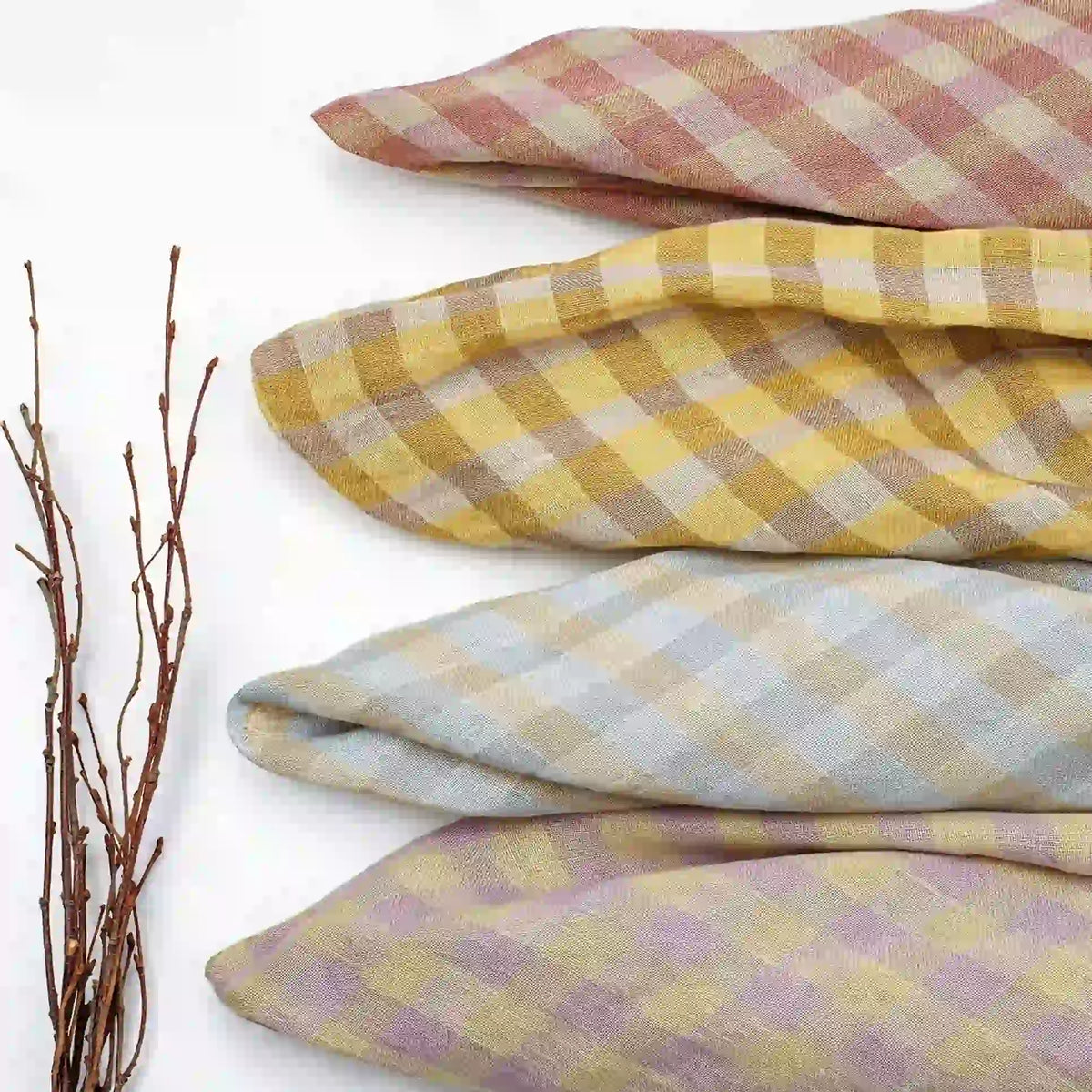 Four folded pieces of the Longan Craft Multicolor Washed Plaid Linen Fabric By The Yard—with vintage checkered designs in pink, yellow, blue, and purple—are arranged horizontally. To the left on the white background, several thin bare twigs are displayed.