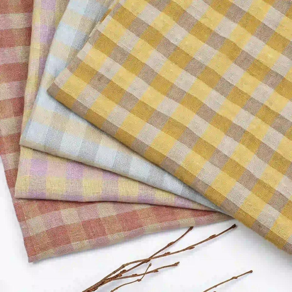 Three folded pieces of Longan Craft’s Multicolor Washed Plaid Linen Fabric by the yard—vintage checkered pure linen in pastel yellow, pink, and blue—are arranged on a white surface with thin brown twigs in the foreground.
