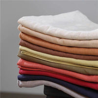 A stack of Longan Craft Multiple Colors Linen Fabric By The Yard—washed cotton linen blend in white, beige, brown, yellow, red, and navy—is neatly arranged on a dark surface against a neutral background.