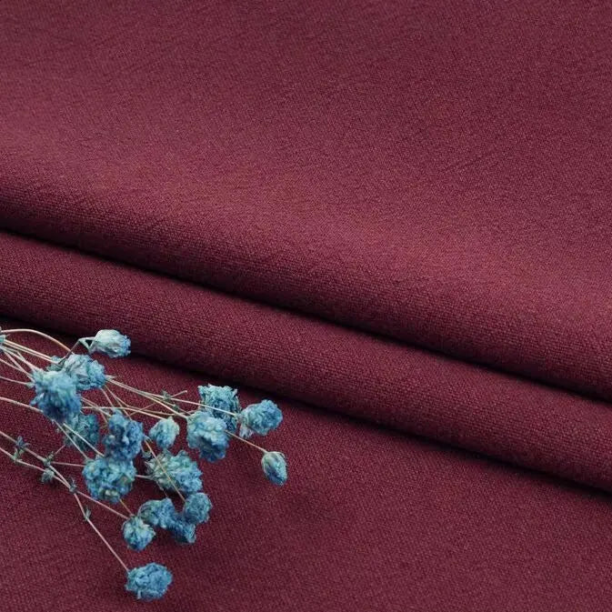 A sprig of small, dried blue flowers lays on neatly folded maroon linen fabric by Longan Craft—washed cotton linen blend by the yard, offering softness, breathability, and durability for apparel and clothing projects.