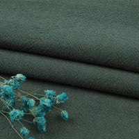 A close-up of Longan Craft’s Multiple Colors Linen Fabric By The Yard, a washed cotton linen blend for apparel, in dark green with a textured surface. Dried blue flowers rest on the solid fabric in the lower left corner.