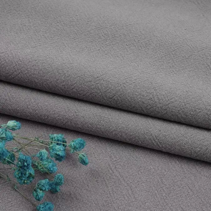 Multiple colors of Longan Craft's washed cotton linen blend fabric by the yard are neatly stacked, showing off their textured durability for apparel, with a small blue dried flower sprig beside them.