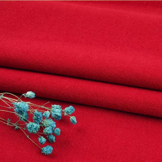 A few light blue dried flowers rest on neatly folded vibrant red linen fabric by the yard from Longan Craft, made of a washed cotton linen blend that's breathable and durable—ideal for apparel.