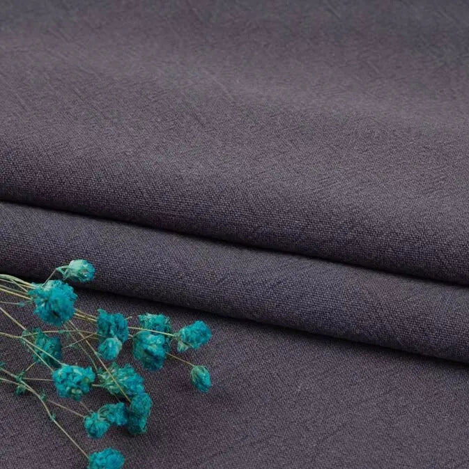 A close-up shows dark gray Longan Craft Multiple Colors Linen Fabric by the yard, a breathable washed cotton linen blend for apparel, folded neatly with a sprig of dried blue flowers on top.