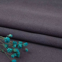 A close-up shows dark gray Longan Craft Multiple Colors Linen Fabric by the yard, a breathable washed cotton linen blend for apparel, folded neatly with a sprig of dried blue flowers on top.