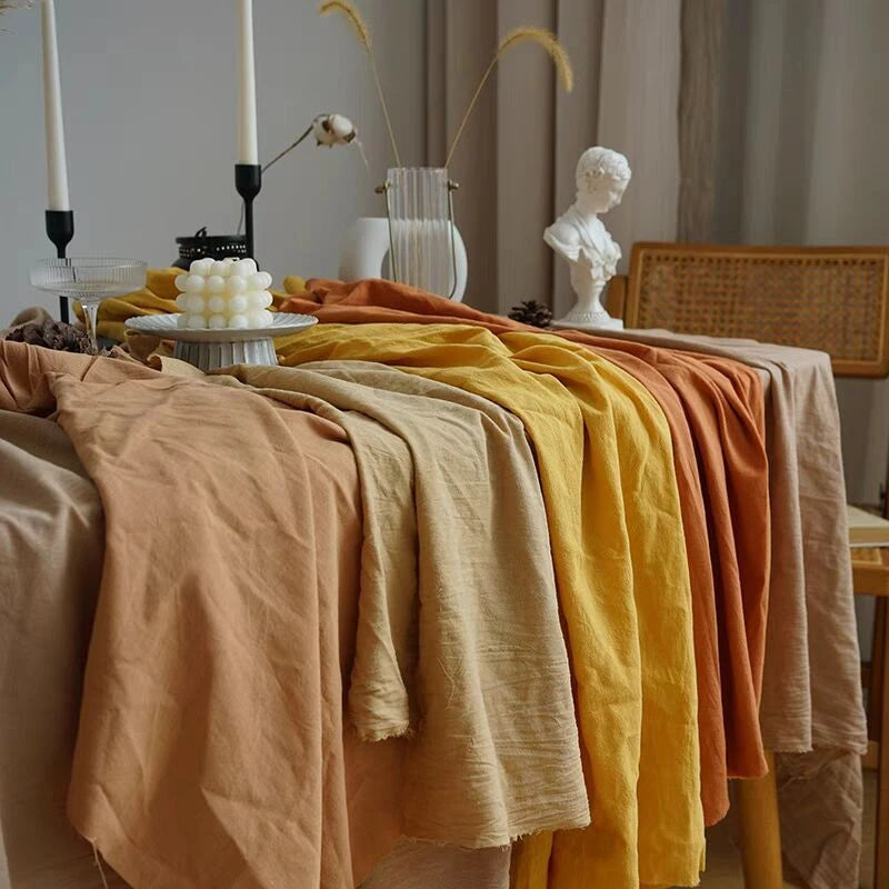 Longan Craft’s Multiple Colors Linen Fabric By The Yard in beige, yellow, and orange drapes over a table near candles, a vase with dried plants, and a white bust for a warm, artistic setting.