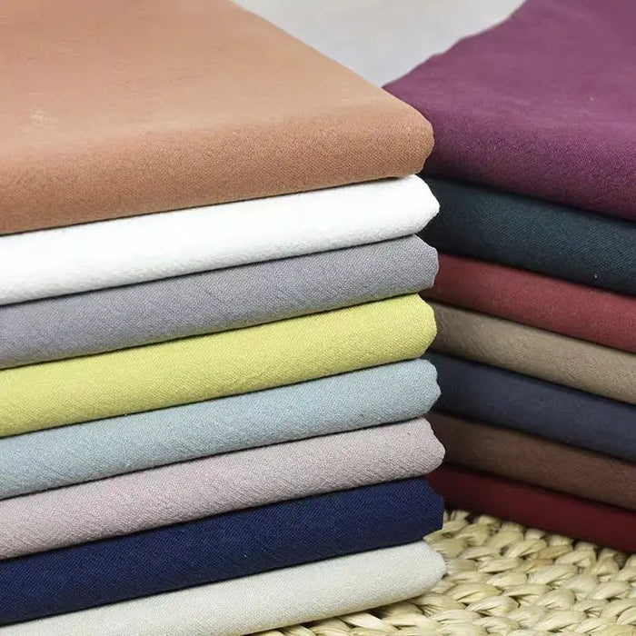 Stacks of Longan Craft’s Multiple Colors Linen Fabric By The Yard—washed cotton linen blend in brown, white, gray, yellow, blue, green, and burgundy—are displayed on a woven mat.