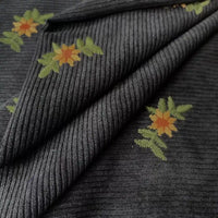 Close-up of Longan Craft’s Thick 8 Wale Polyester Corduroy Fabric by the yard, featuring ribbed dark corduroy embroidered with yellow and orange floral designs and green leaves. The folded fall upholstery textile is ideal for apparel.