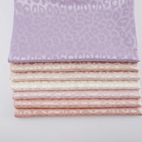 A stack of pastel fabrics—lavender, cream, and light pink—made from Longan Craft's Leopard Jacquard Satin Fabric by the yard, featuring a subtle leopard print on soft, smooth polyester satin for DIY shirts.