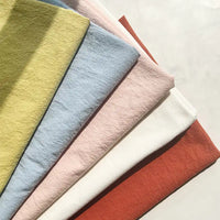 Six pieces of Longan Craft 100% Cotton Crepe Fabric by the yard, in a selection of pastel and earthy shades—yellow, blue, light pink, cream, and rust—are neatly layered atop a light surface. These pure color washed crepe fabrics are perfect for breathable casual wear clothing.