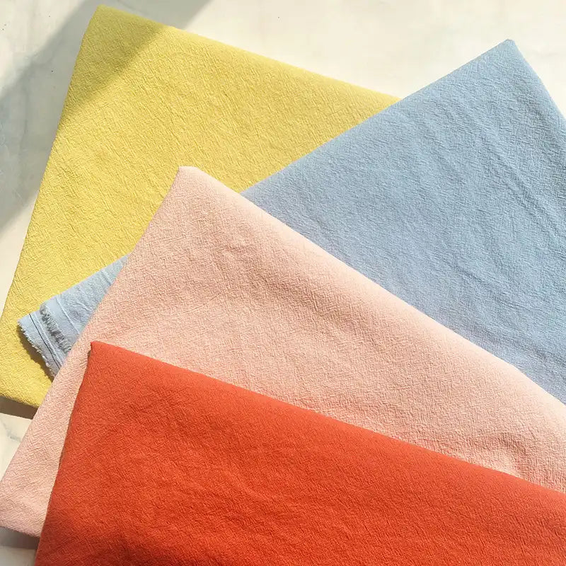 Four pieces of Longan Craft’s 100% Cotton Crepe Fabric By The Yard—ideal for those wanting pure color washed crepe material for casual clothing—are neatly stacked in pastel yellow, light blue, pale pink, and bright orange on a light surface, with sunlight casting soft shadows.