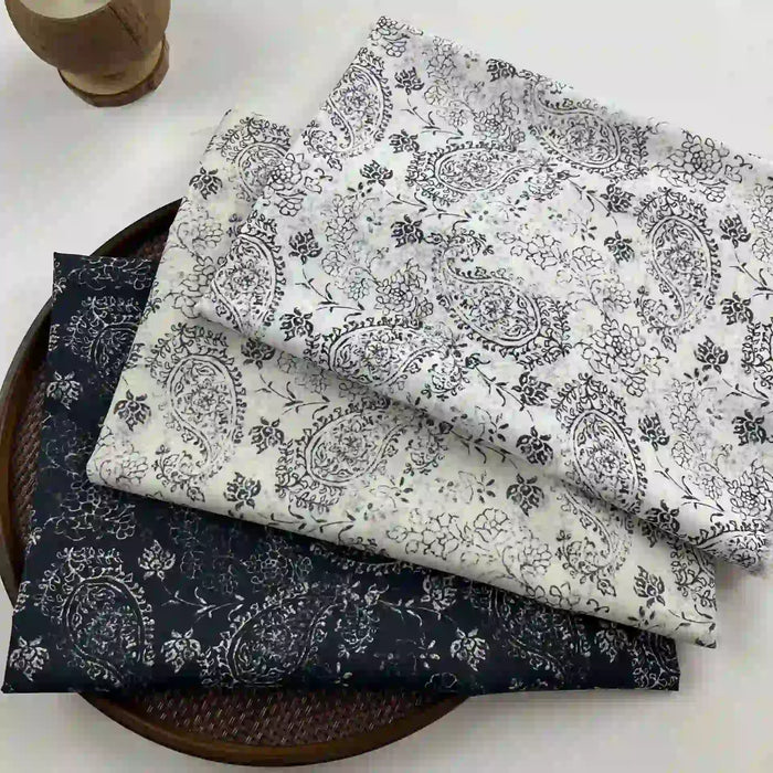 Three folded pieces of Longan Craft Paisley Printed Voile Fabric By The Yard, pure cotton with black and white floral and paisley patterns on white, cream, and black backgrounds, are stacked on a round tray. Ideal for shirts.