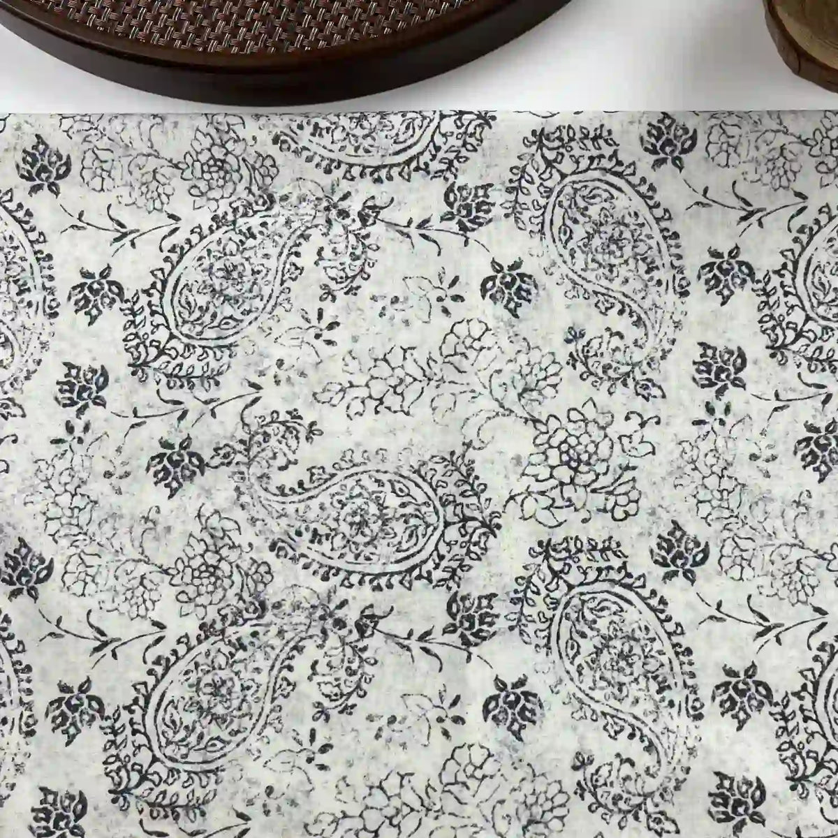 White surface features the Paisley Printed Voile Fabric by Longan Craft: pure cotton with black floral and paisley motifs. Part of a woven dark brown circular object appears at the top edge. Ideal for shirts, sold by the yard.